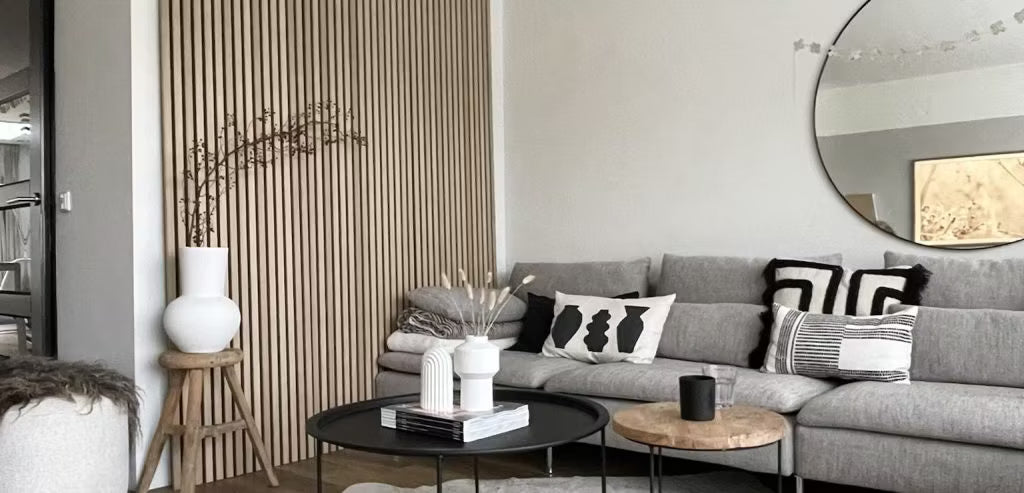 Modern living room featuring a gray sofa, abstract pillows, black coffee tables, decorative vases, and an Akupanel wood acoustic slatwall with a round mirror.